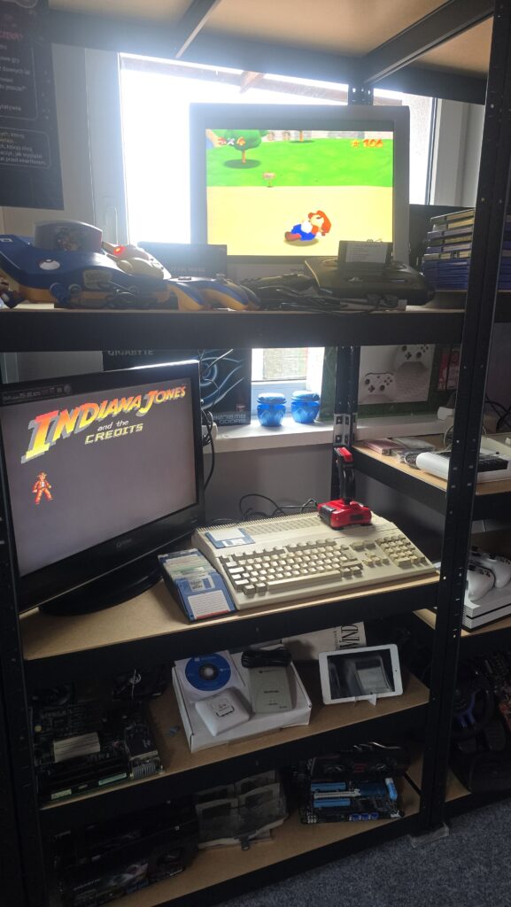 Regał ze stanowiskami do gier retro Amiga, Atari, Sega, Nintendo, monitorami wyświetlającymi klasyczne gry, joystickiem i starym komputerem z klawiaturą